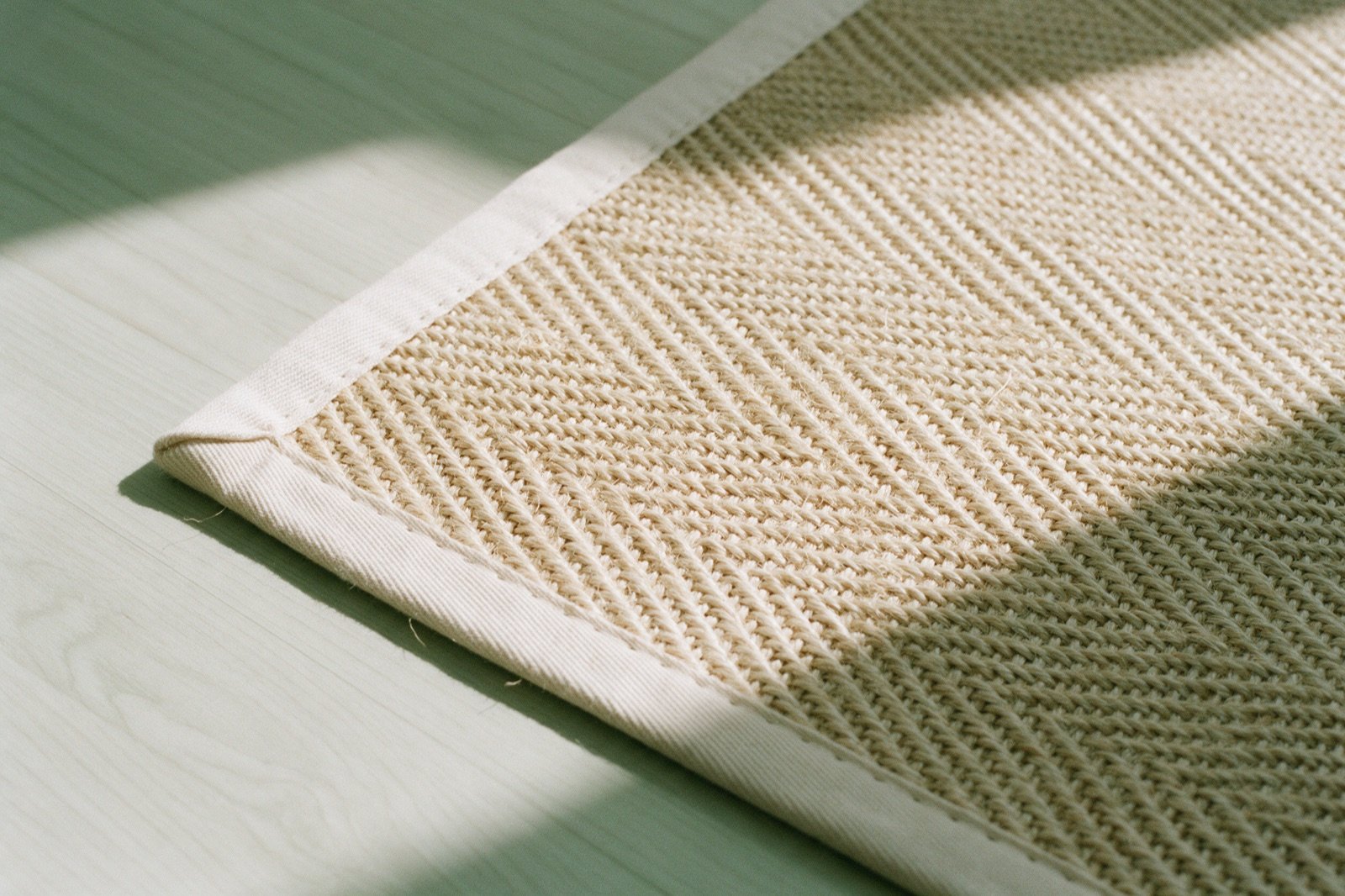 Detalhe de um tapete de sisal bege com trama grossa em espinha e acabamento de cadarço na borda, sobre piso de madeira